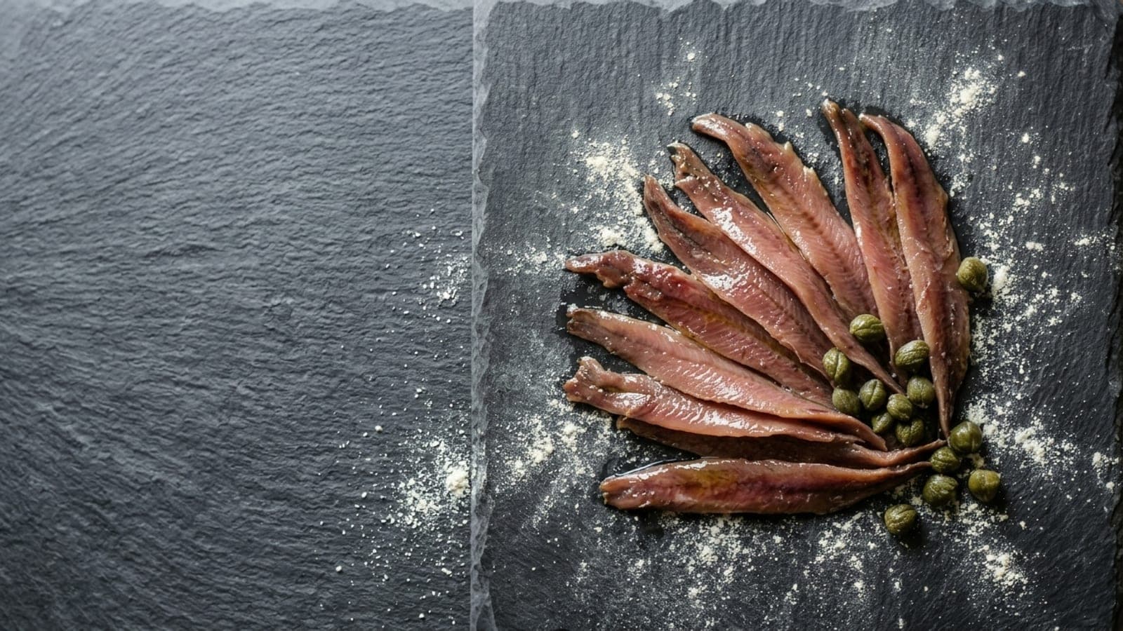 Glistening oil-packed anchovy fillets fanned out with a scatter of rinsed capers between them, on dark slate