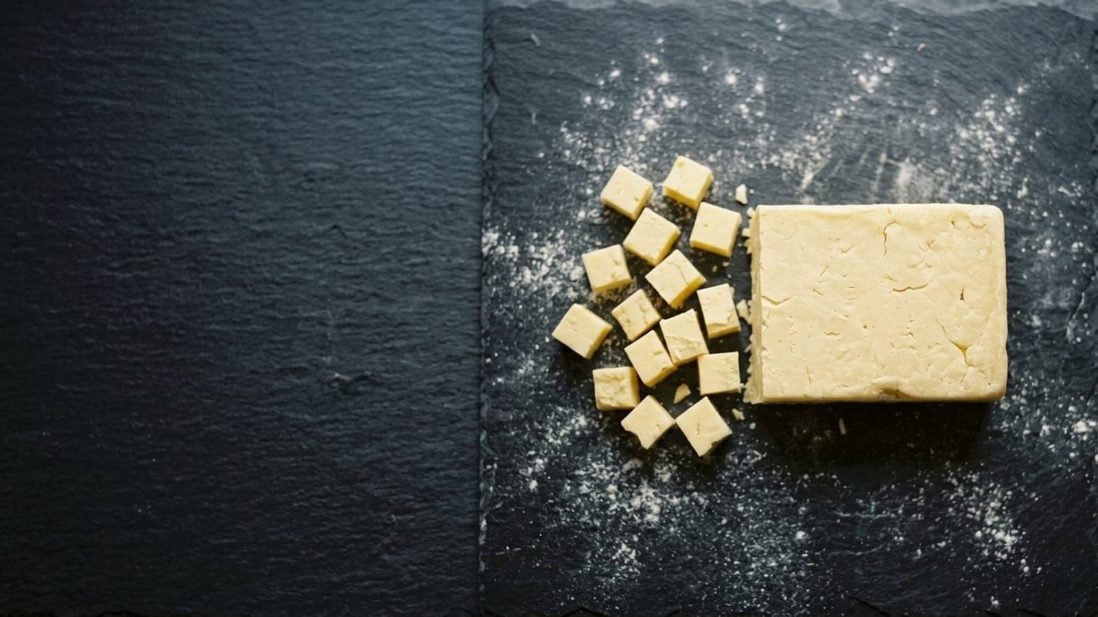 A block of pale brick cheese partly cut into small cubes on dark slate