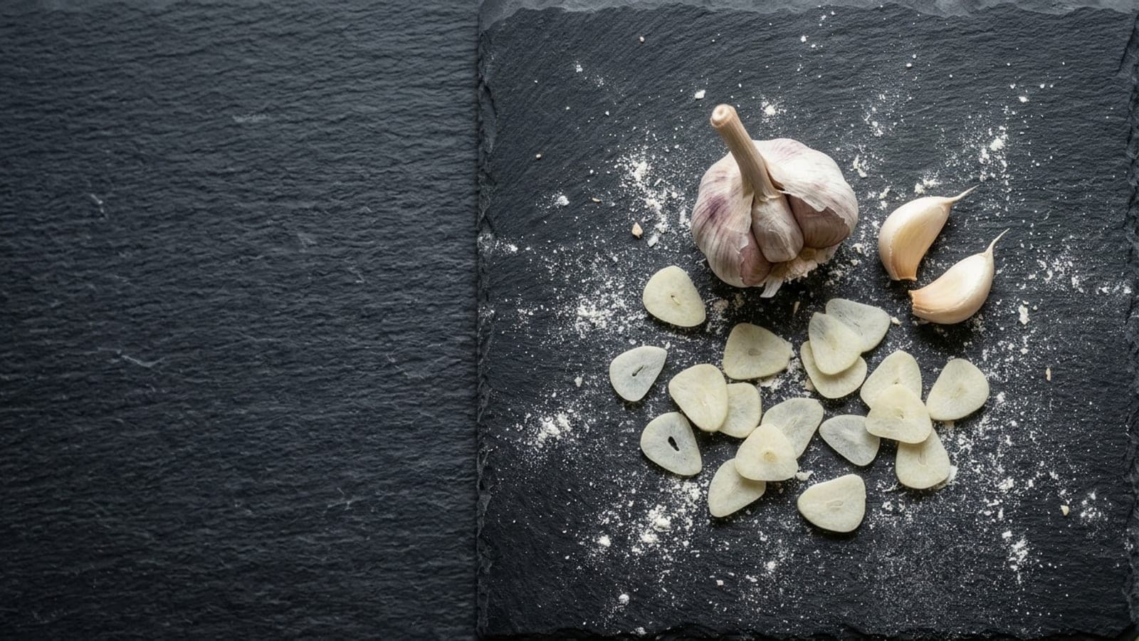 A head of garlic with peeled cloves and paper-thin slices scattered on dark slate