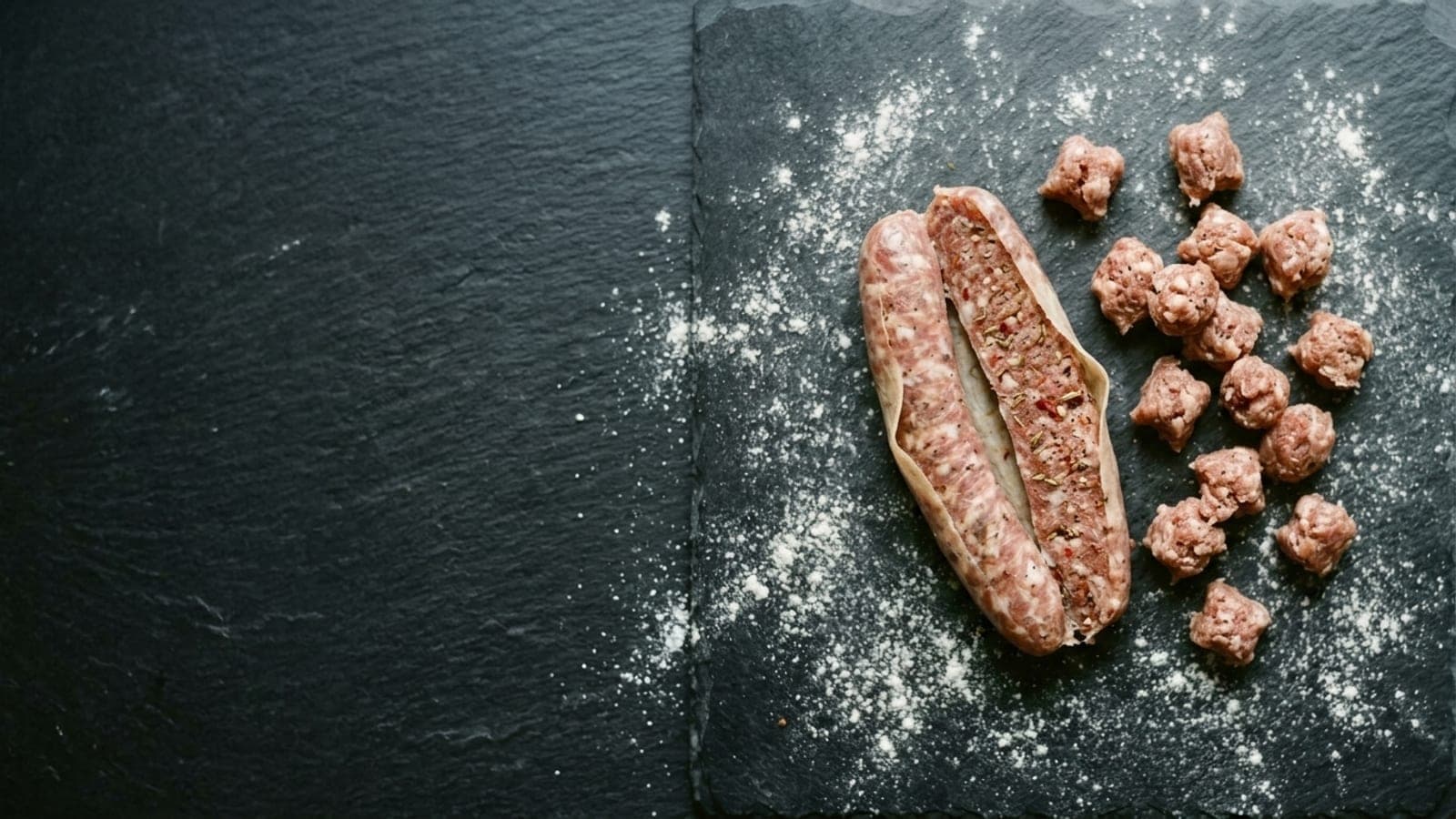 A raw Italian sausage split open with the casing peeled back and pinched nuggets of sausage meat on dark slate