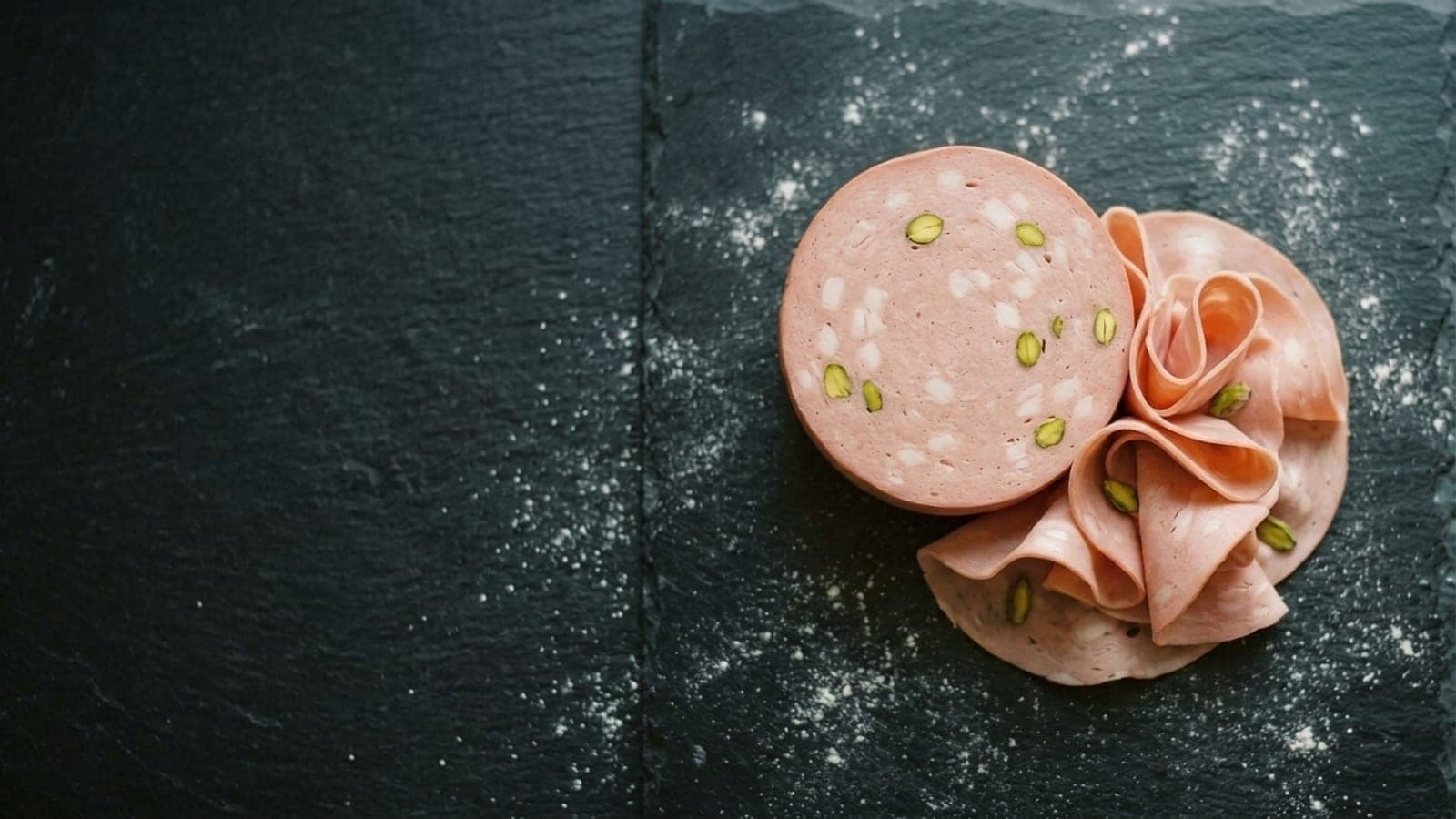 A thick round of pistachio-studded mortadella, partly sliced, with thin folded slices draped beside it on dark slate