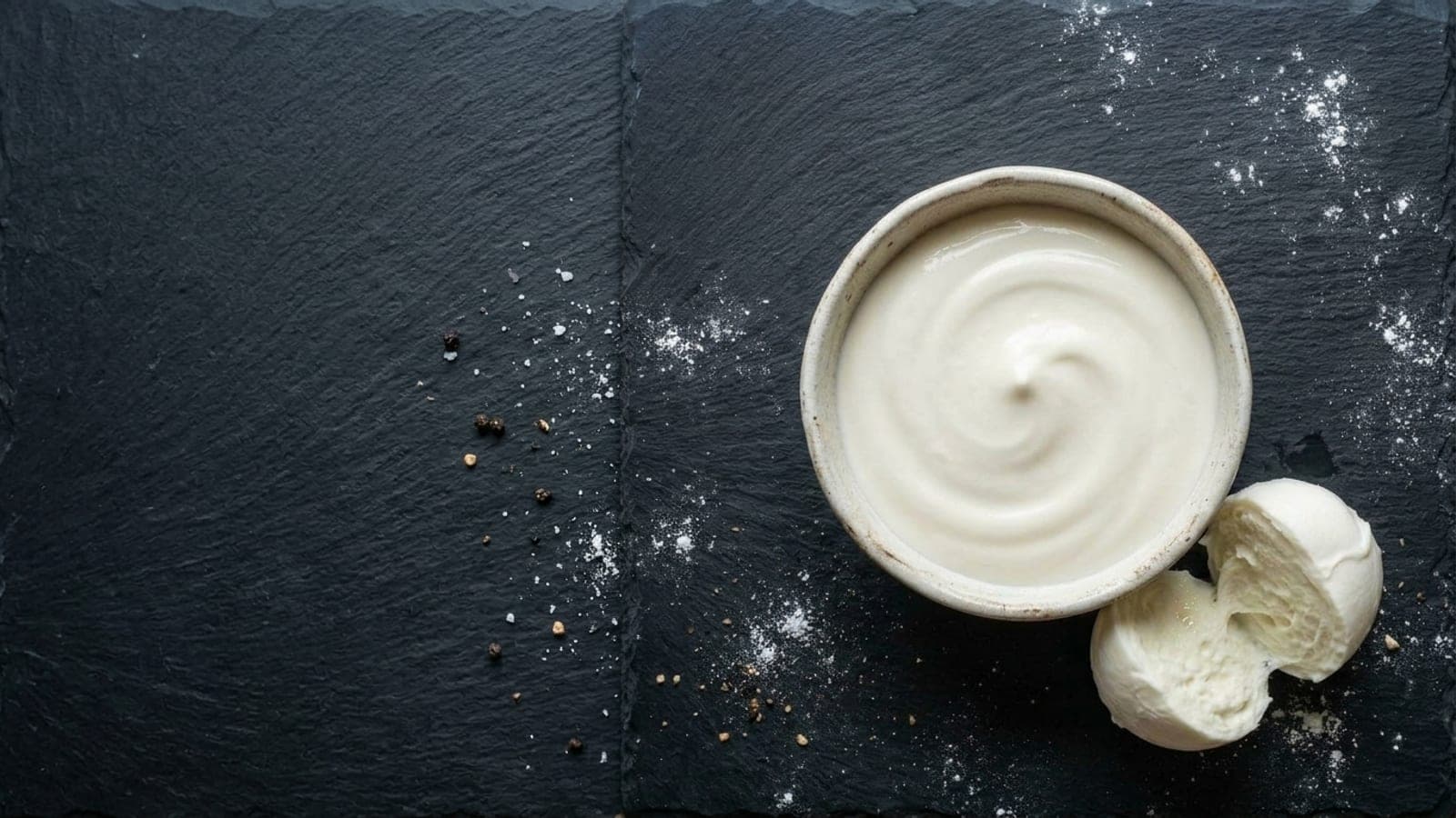 A bowl of thick, smooth white mozzarella cream with a torn ball of fresh mozzarella beside it, on dark slate