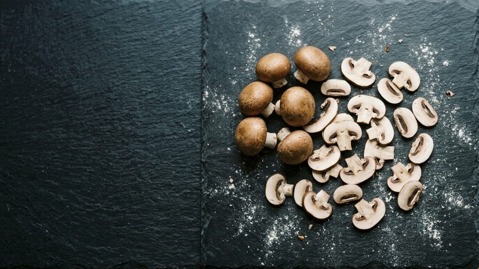 Whole cremini mushrooms with thin raw slices lying flat among them on dark slate