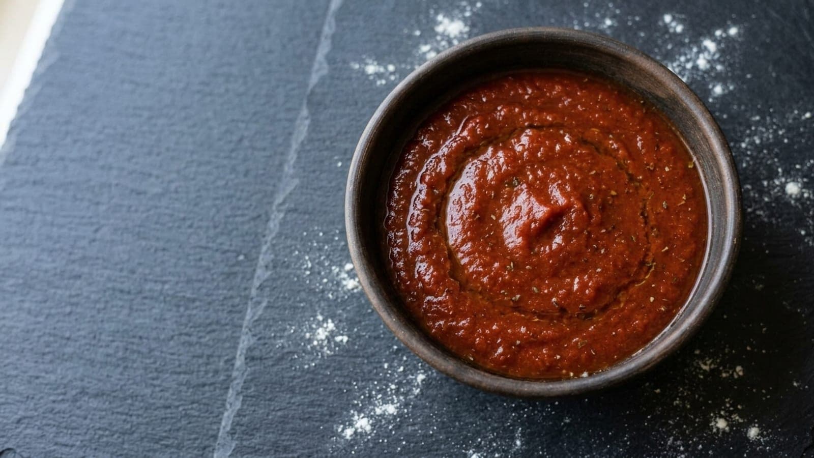 A bowl of smooth New York pizza sauce, deep red with an olive-oil sheen and flecks of oregano, on dark slate