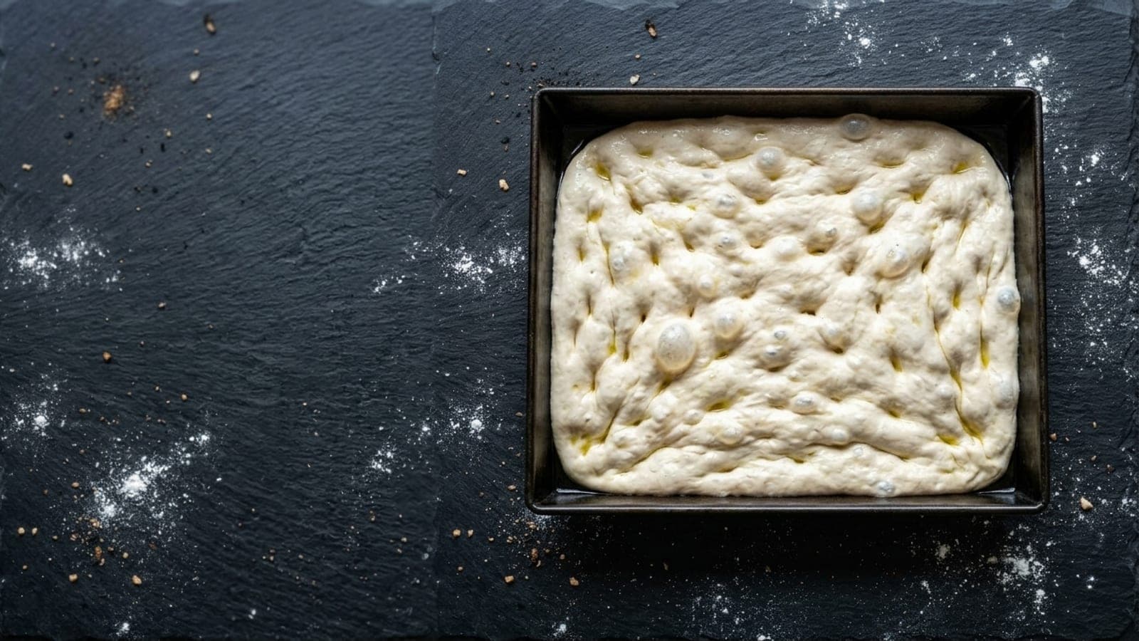 Raw pizza dough dimpled into a rectangular steel pan on a floured dark slate counter.