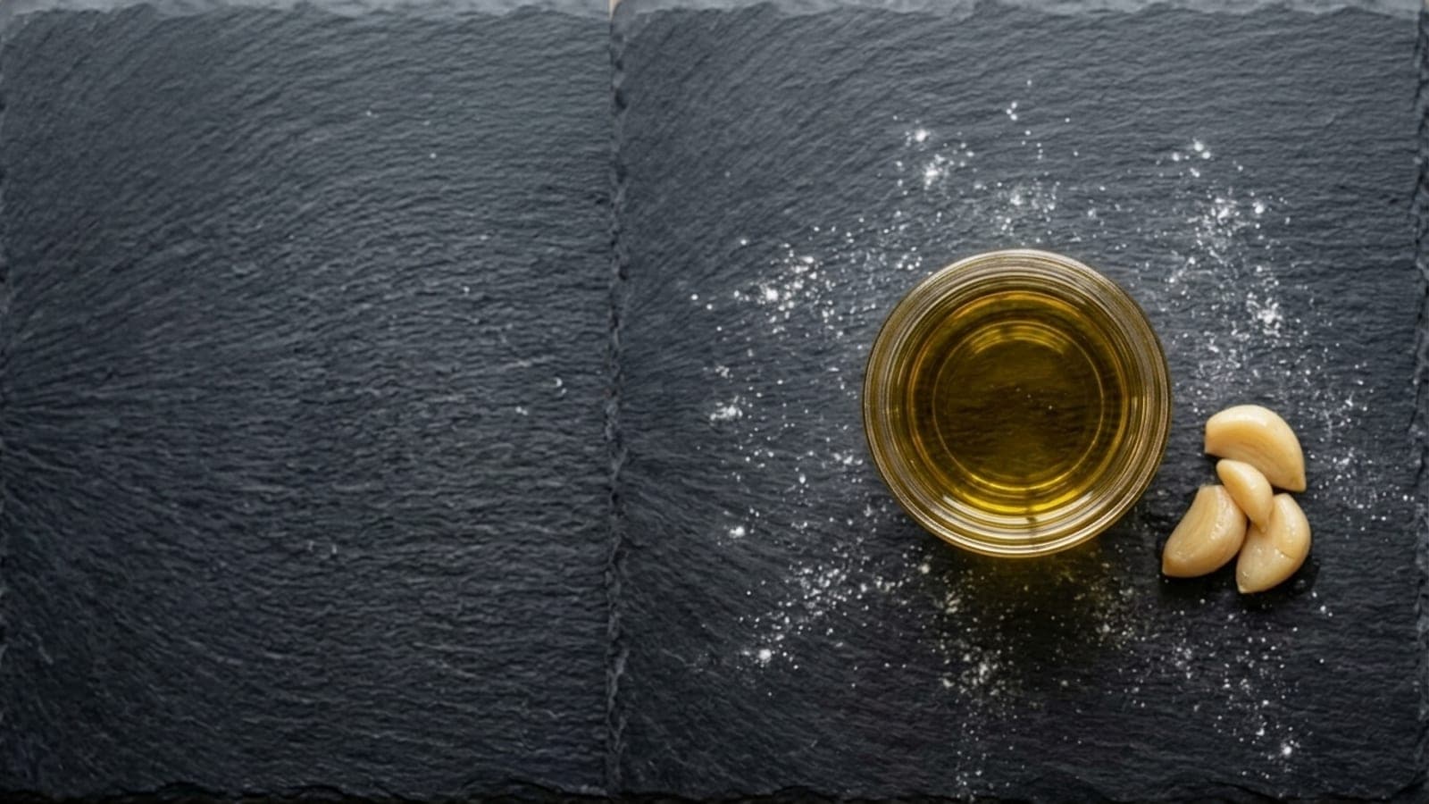 A small glass jar of golden garlic oil, strained clear, with softened cloves beside it on dark slate