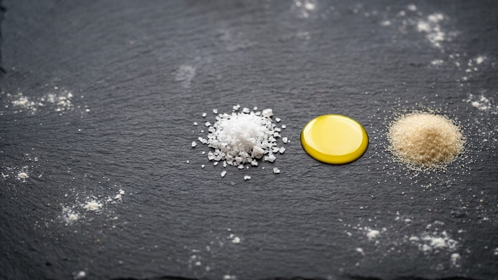 Small heaps of flaky salt, a pool of olive oil and fine sugar on a dark slate counter.