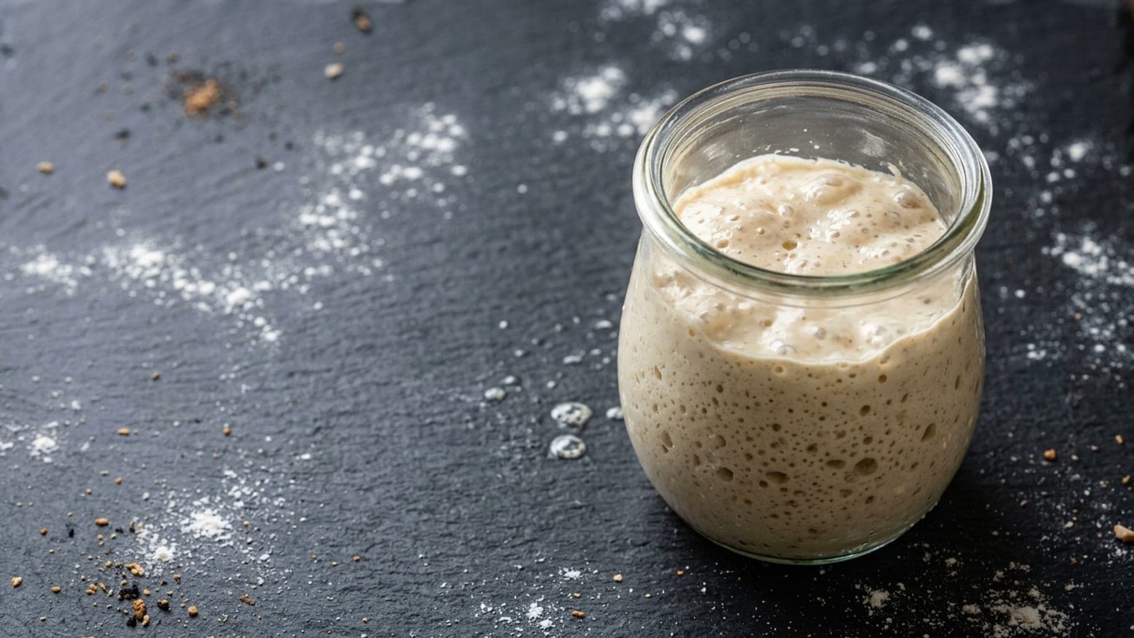 A jar of active, bubbling sourdough starter on a floured dark slate counter.