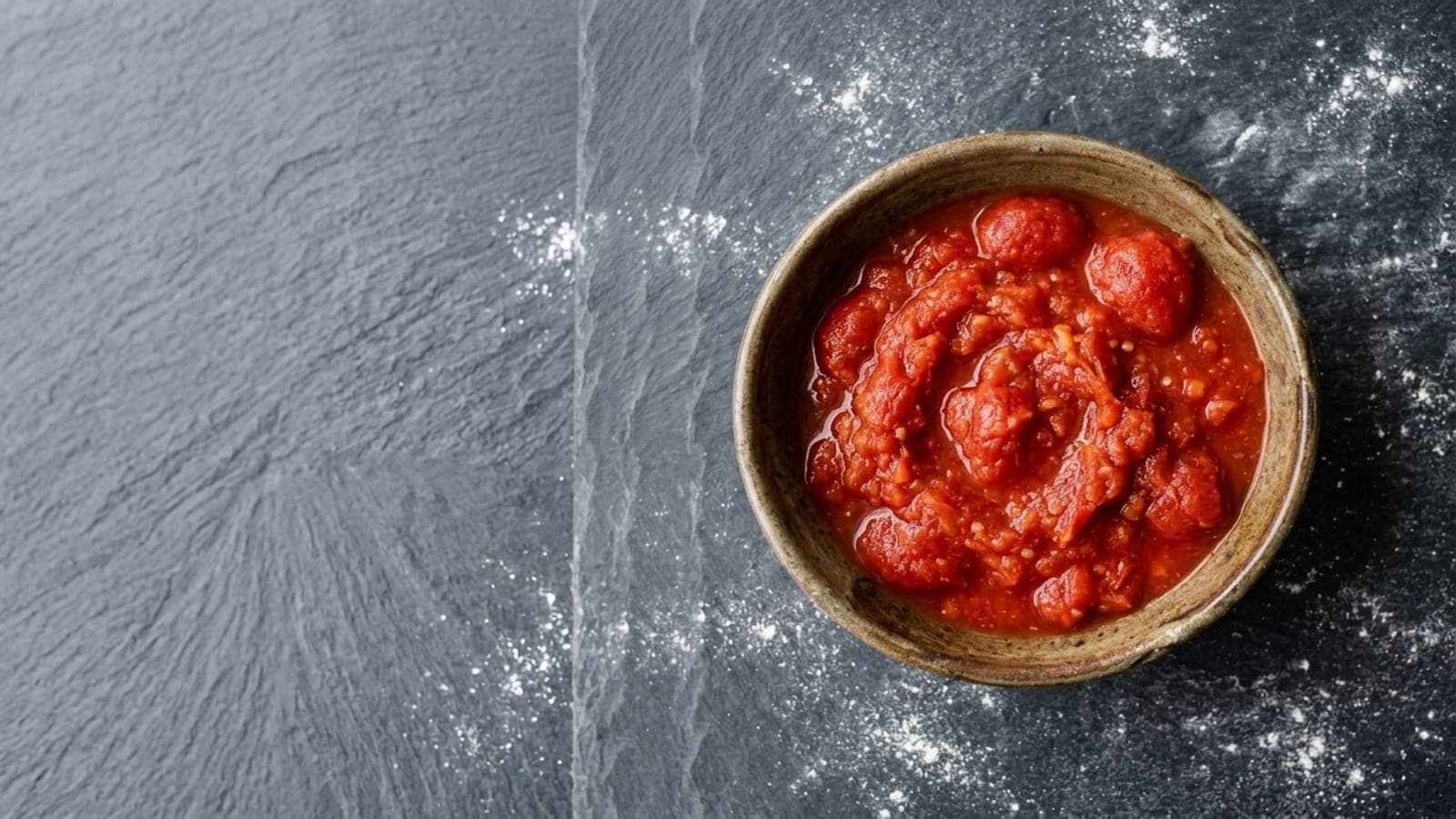 A bowl of raw tomato sauce, hand-crushed and pulpy with pieces of tomato still visible, on dark slate