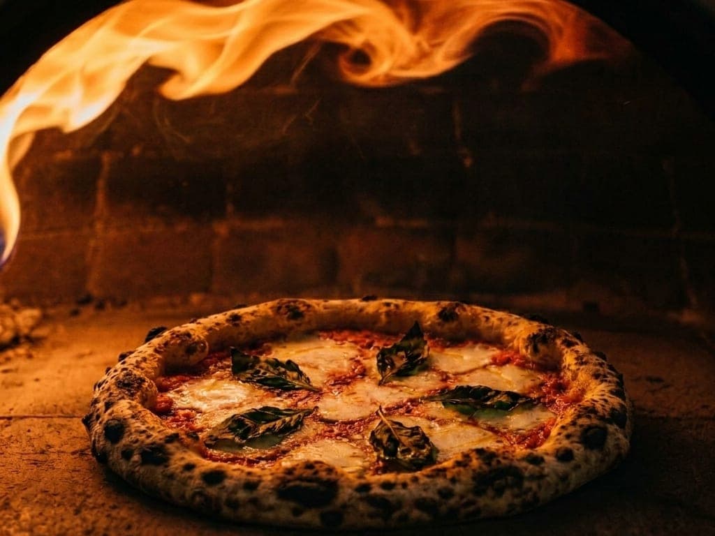 A Neapolitan pizza baking on the glowing stone floor of a wood-fired oven, orange flame rolling across the brick dome above it and the rim charring into leopard spots